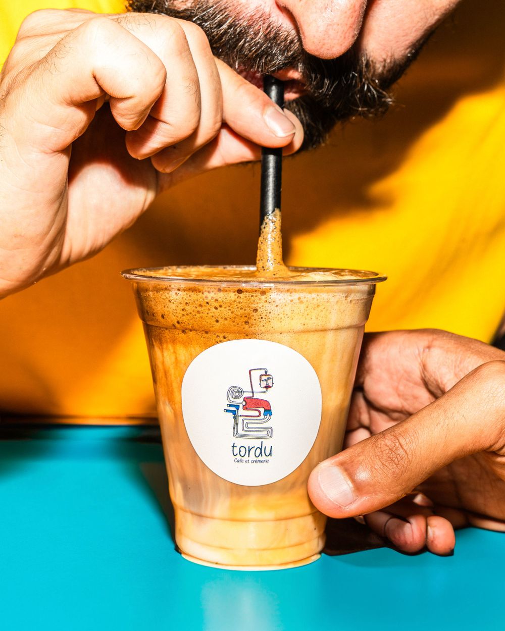 Person stirring iced coffee drink with branded cup logo against colorful background. Mtlshutter photography