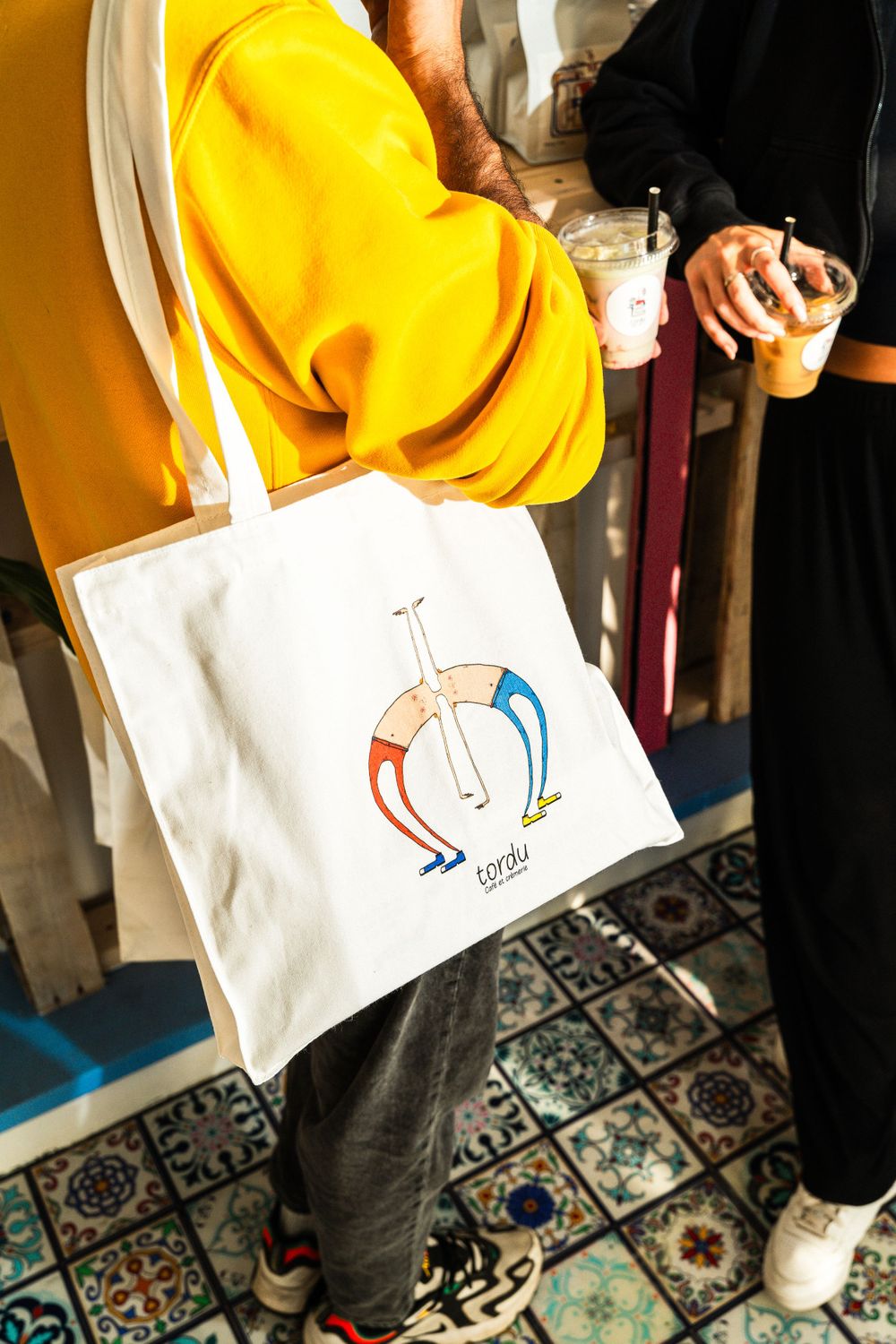 Person wearing yellow sweatshirt holding white canvas tote bag with artistic logo design. Mtlshutter photography