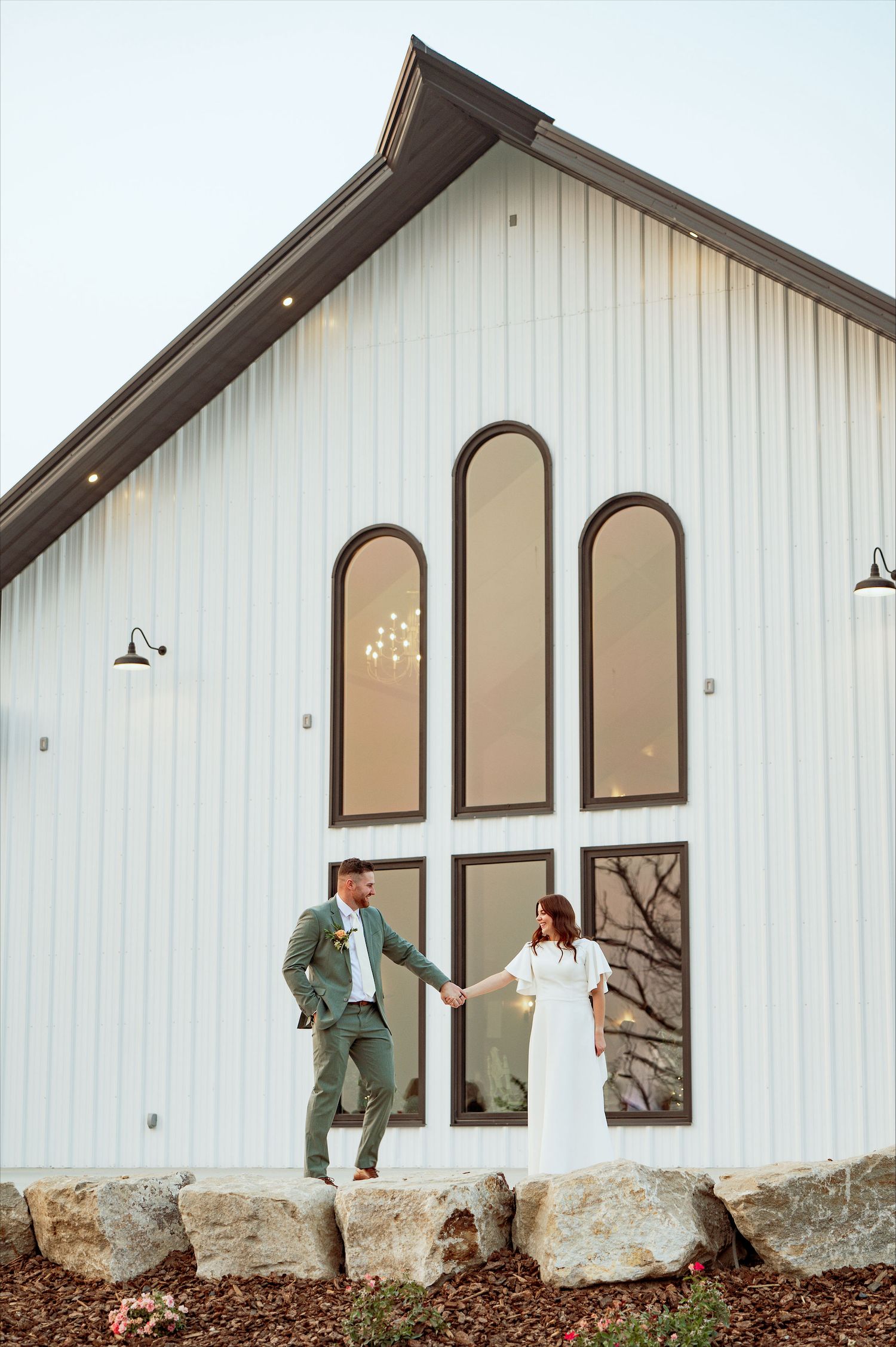 Two people in wedding attire stand outside a modern white barn-style venue with arched windows and stone foundation.