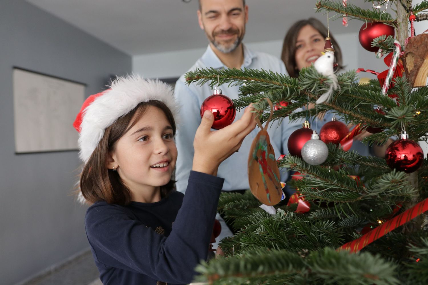 Young girl placing red ornament on Christmas tree while decorating with family.
family photographer