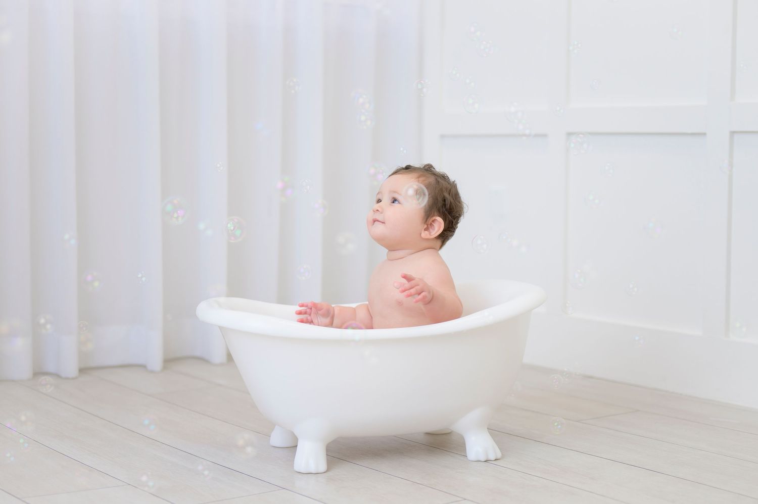 A happy baby sits in a white clawfoot bathtub against a light backdrop during bath time looking up at bubbles.