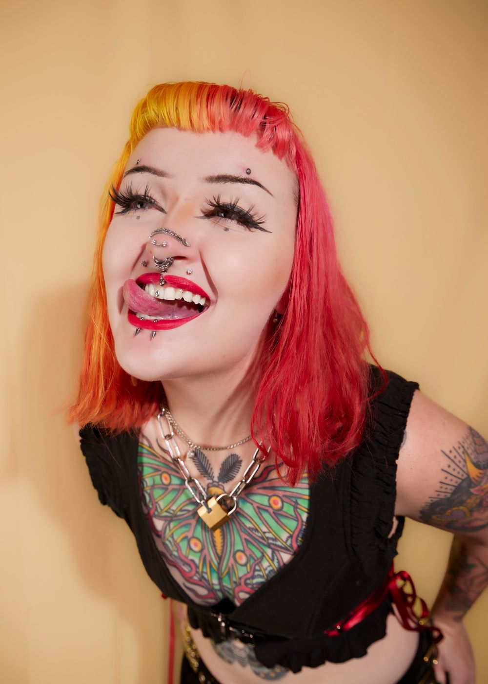 Portrait of a model with tattoos and colourful hair sticking her tongue out against a yellow backdrop