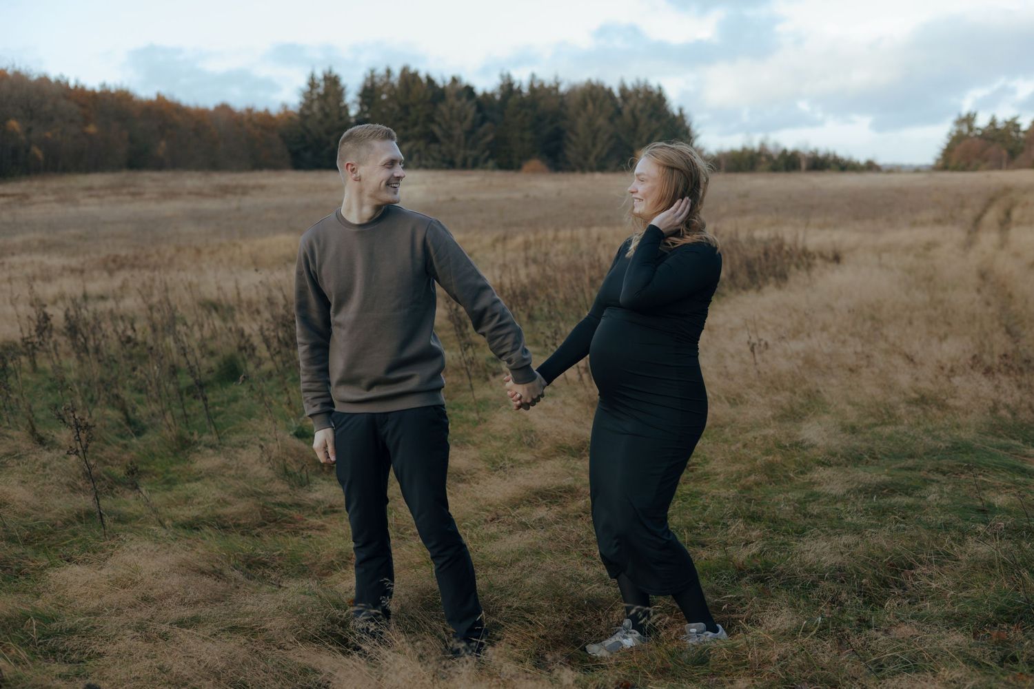 A couple holds hands while walking together in a golden autumn field with forest in the background.