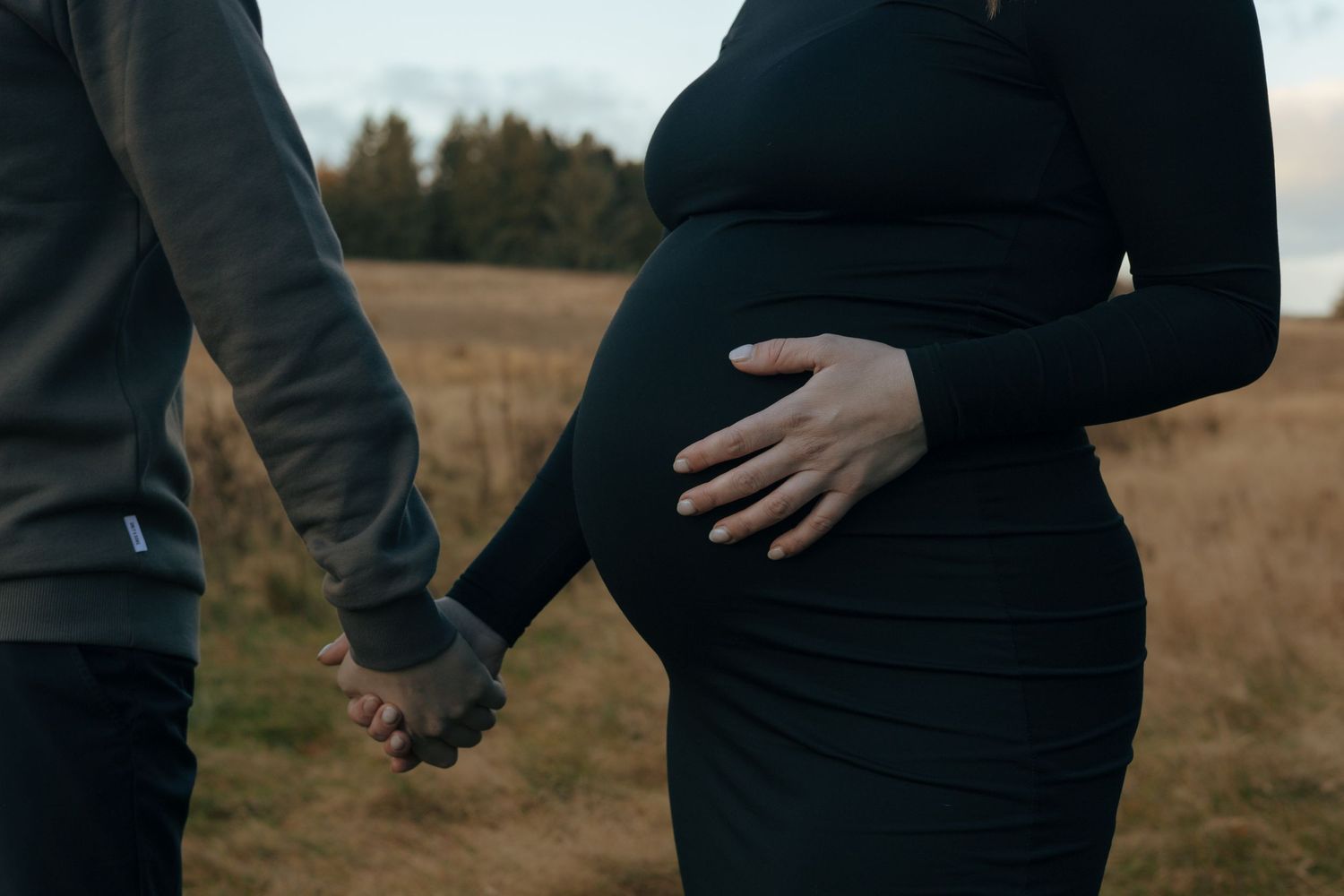 Close-up of a couple holding hands with a pregnant belly visible against an autumn landscape.