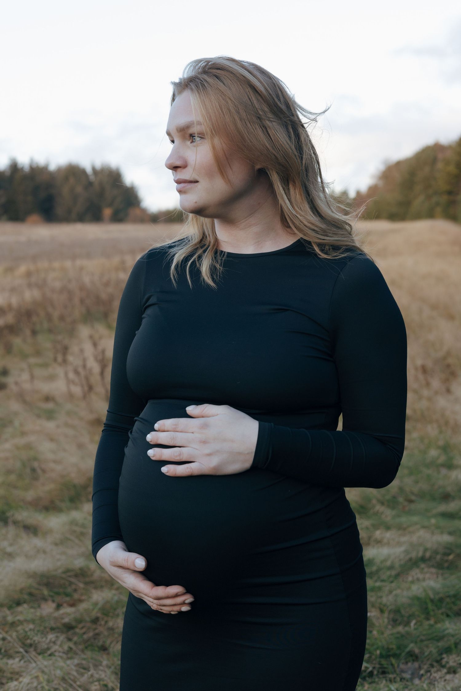 Expectant mother in black dress stands in autumn field during maternity photoshoot at sunset.
