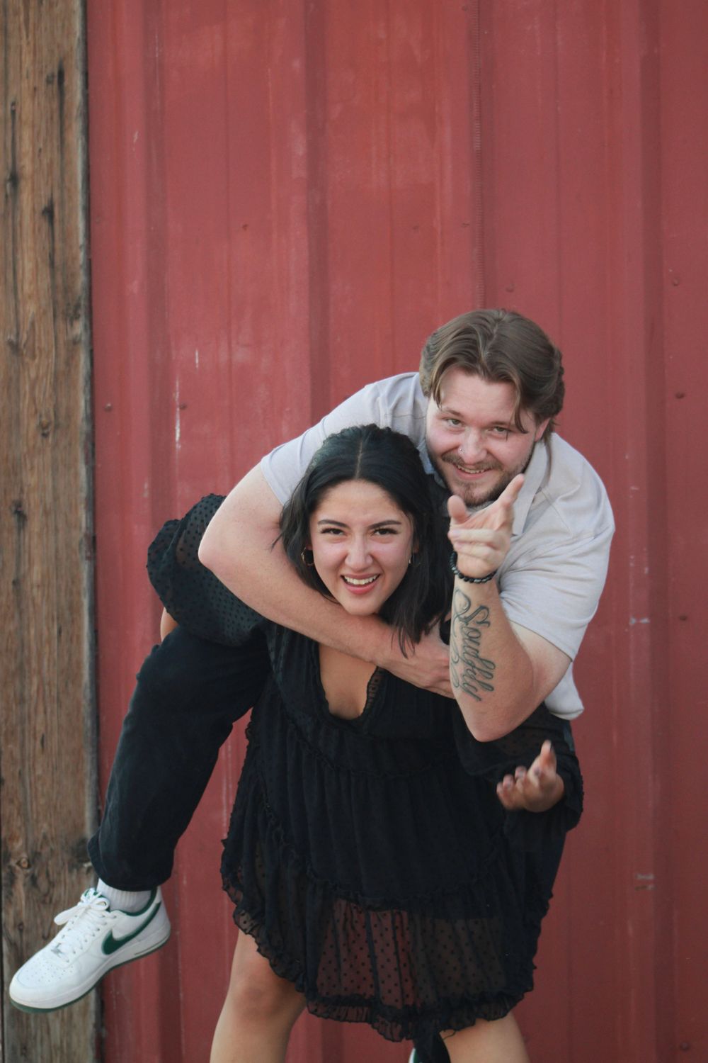 Couple poses playfully with piggyback ride in front of red barn wall.