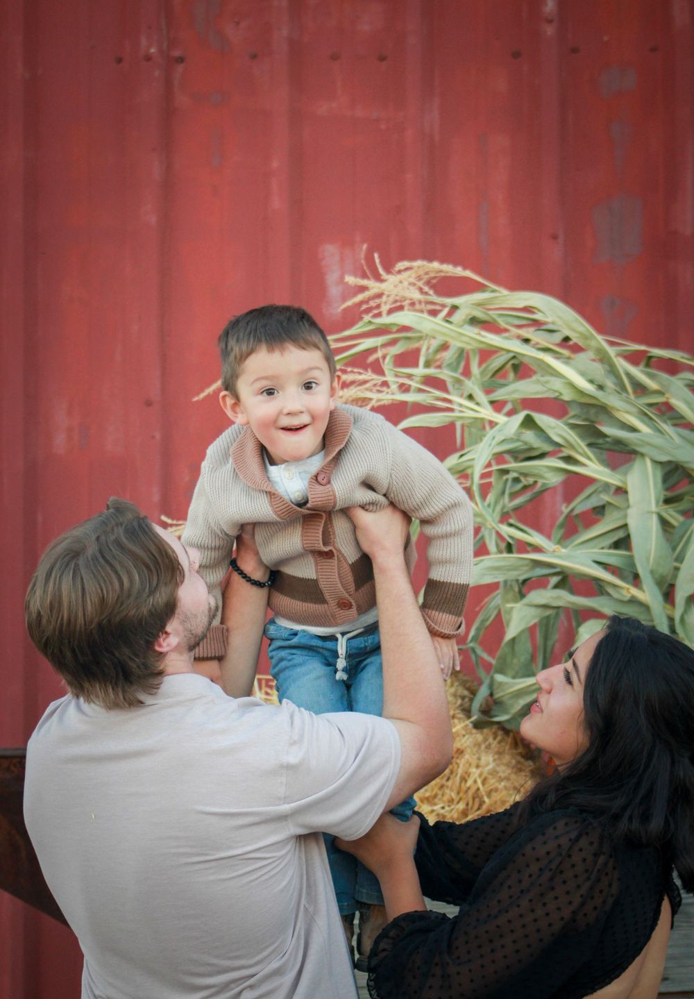 Parents lift child in air during autumn photoshoot by red barn.