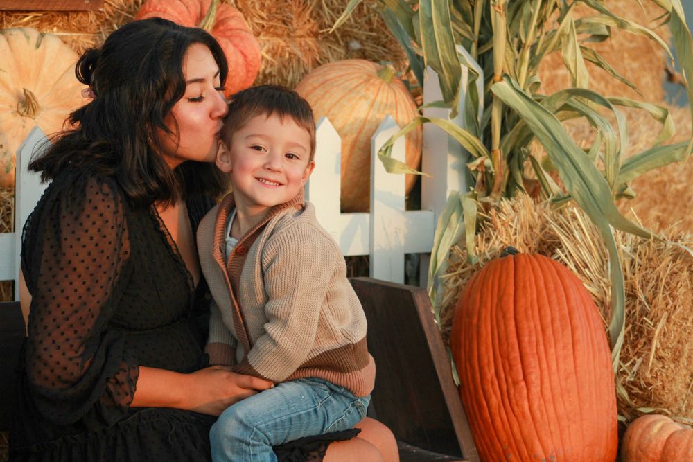 Mother and child share an intimate moment at a fall pumpkin patch with hay bales and white fence.