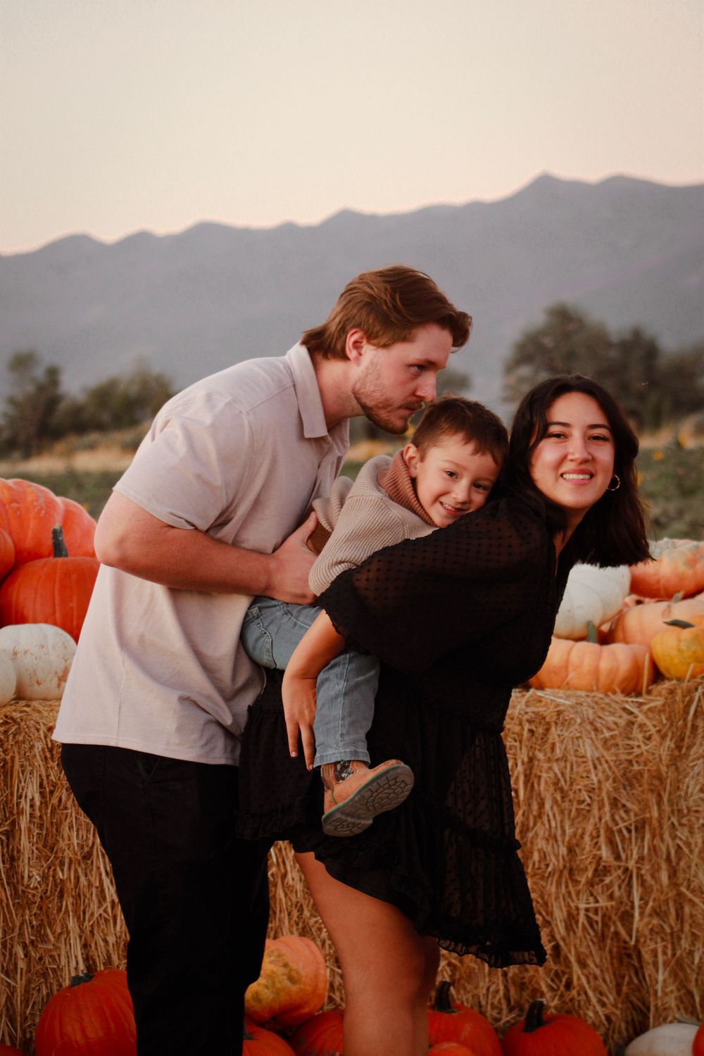 Family of three poses playfully at pumpkin patch during sunset with mountains in background.