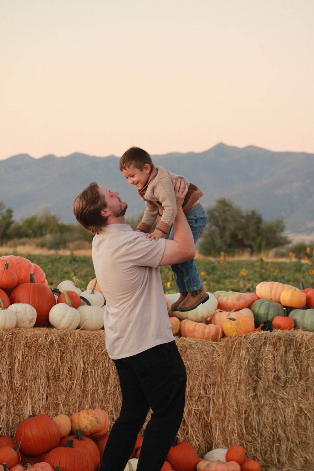Father lifts young son in air at pumpkin patch with hay bales and mountains at sunset.