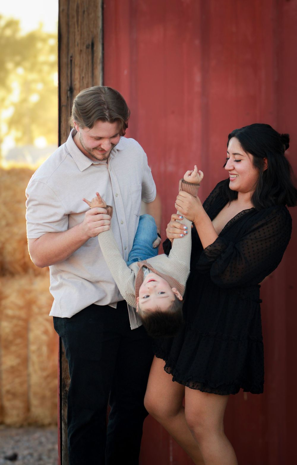 Family shares playful moment holding child upside down by red barn.