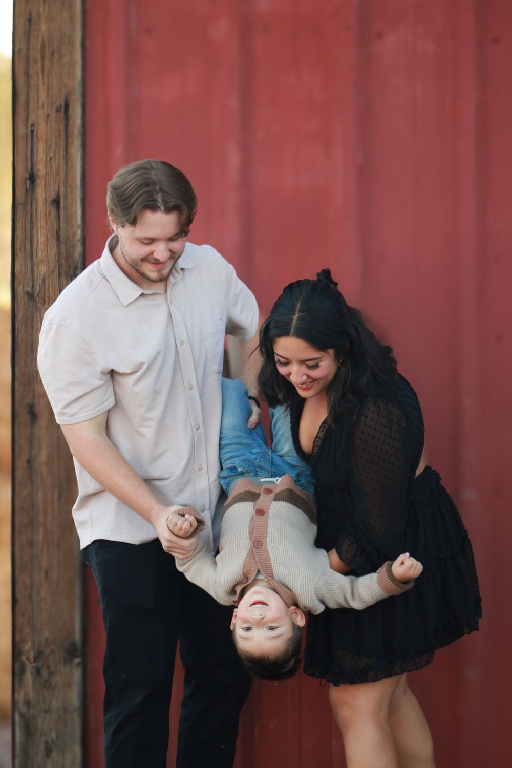 Playful portrait of parents holding child upside down against red barn wall.