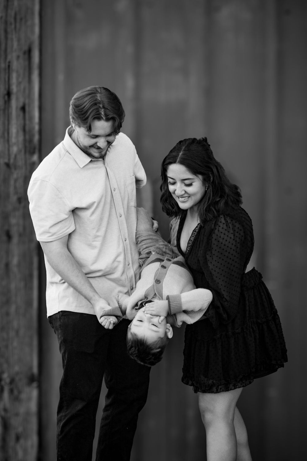 Black and white photo of parents playing with child against rustic backdrop.