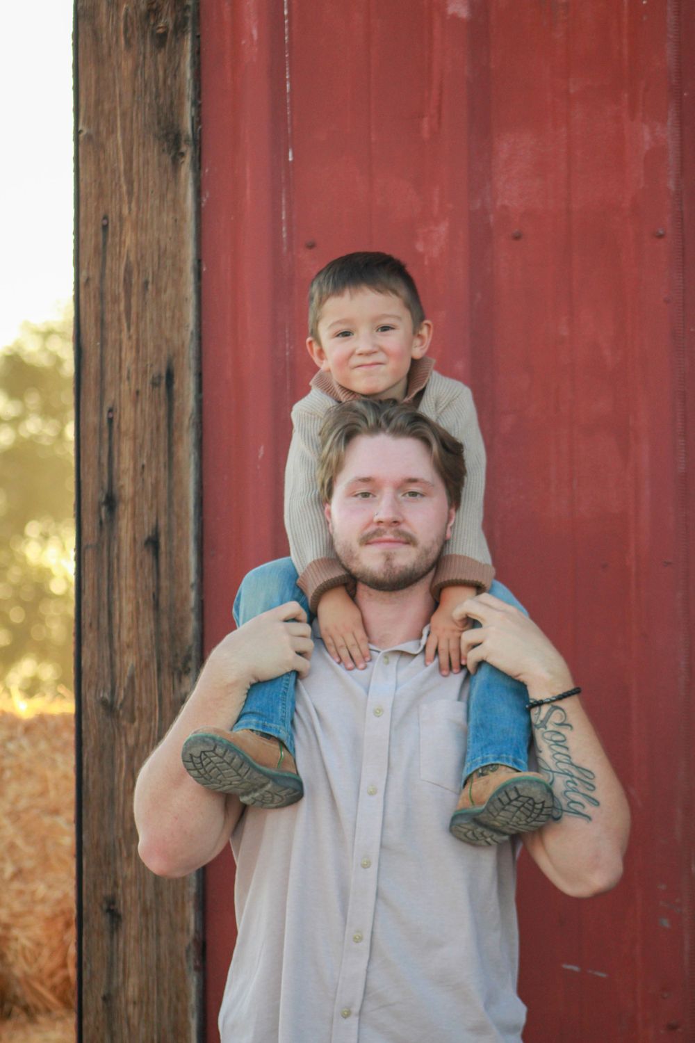 Young child sits on shoulders of person in front of red barn wall.