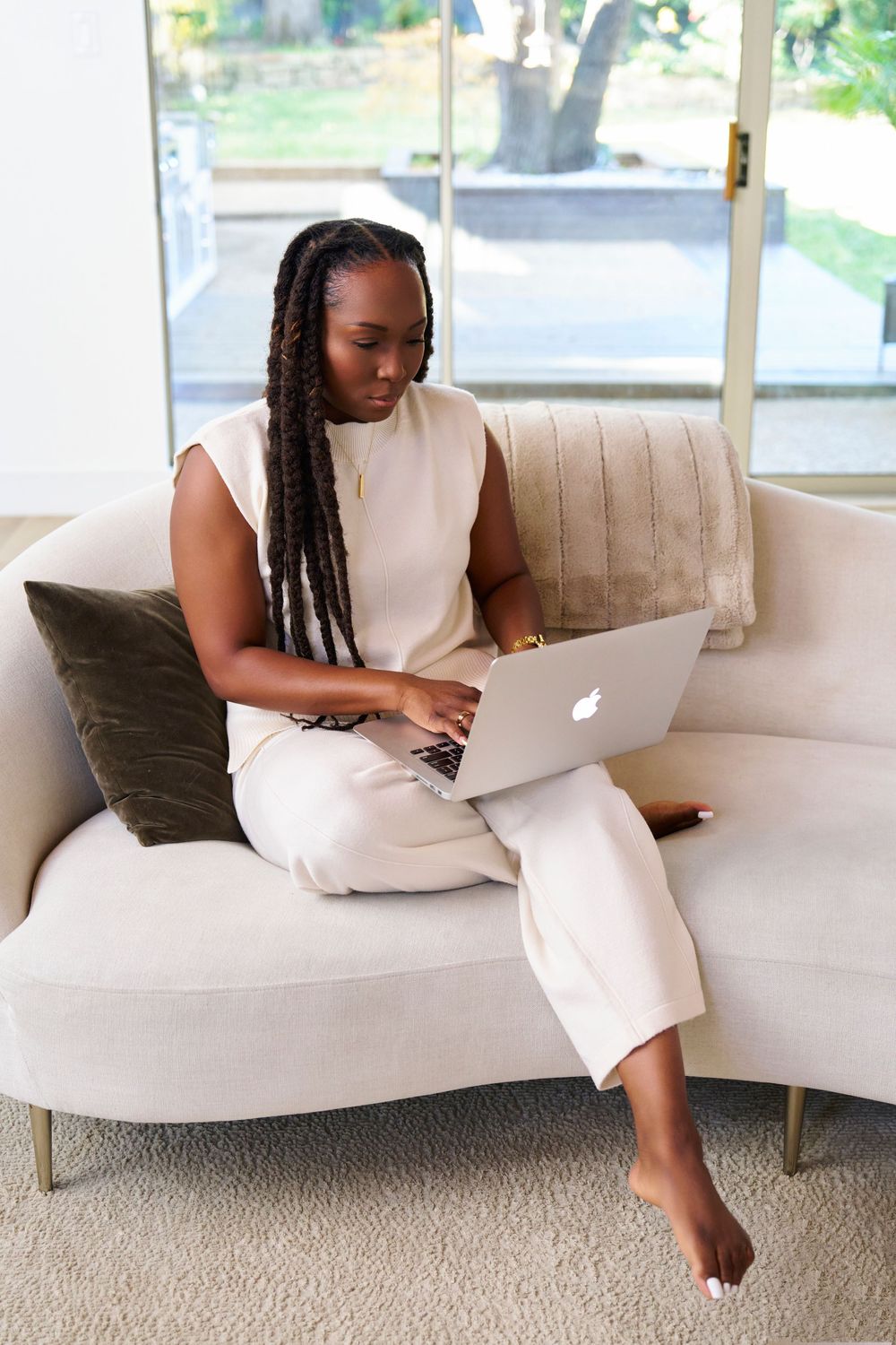 Person in white dress relaxes on beige couch while working on MacBook laptop.