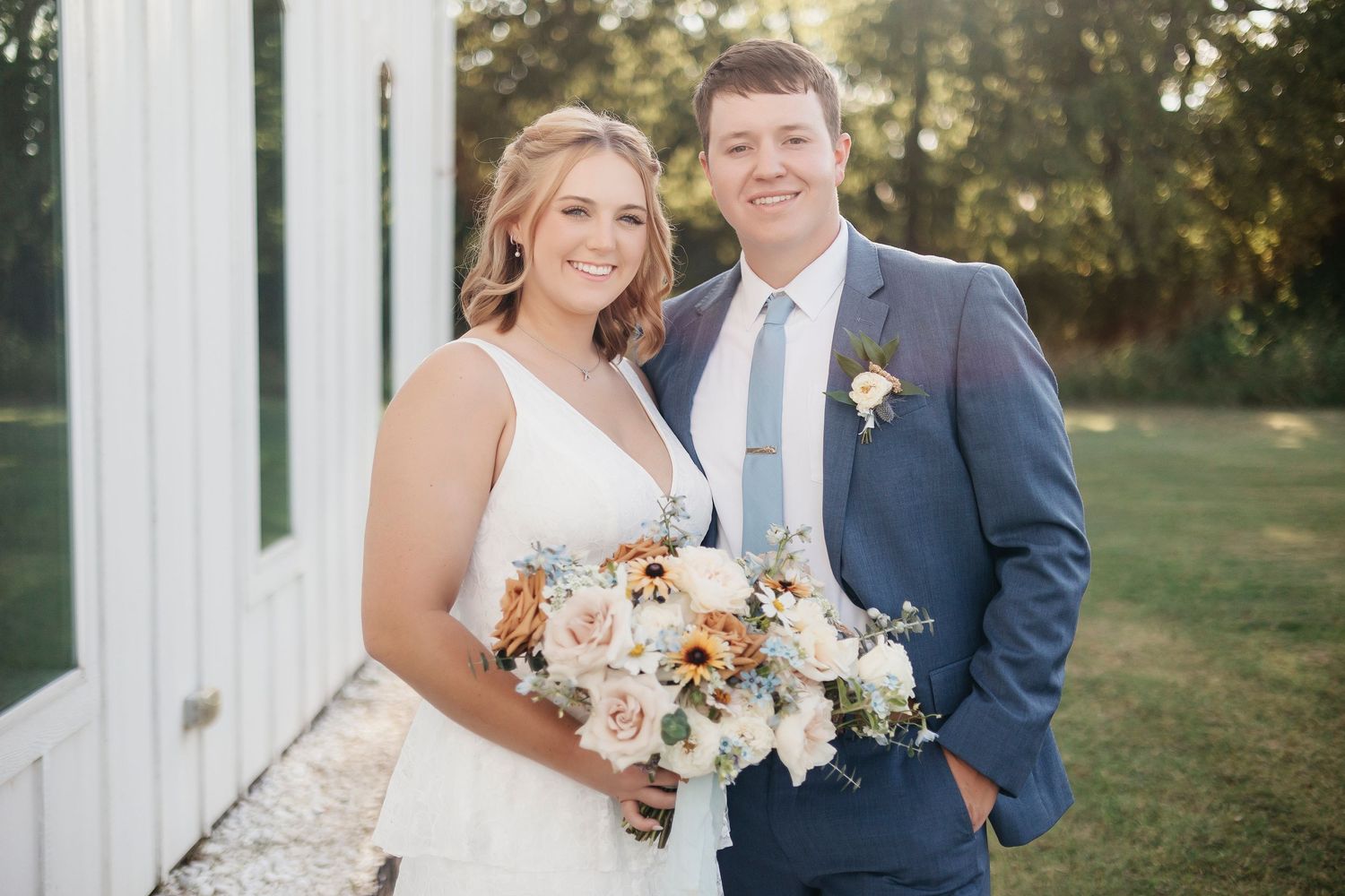Newlywed couple poses outdoors holding peach and white flower bouquet against white building.