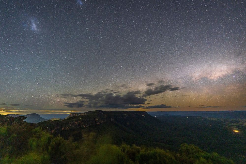 Stunning night sky with Milky Way galaxy and Magellanic Clouds over mountain landscape with glowing horizon.