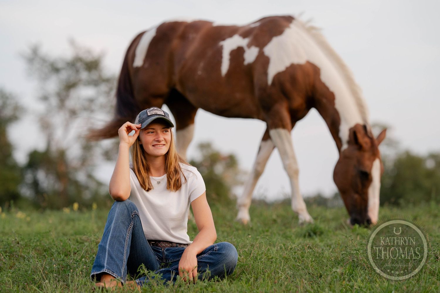 Brown and white horse grazes on hillside while companion in baseball cap sits in foreground.