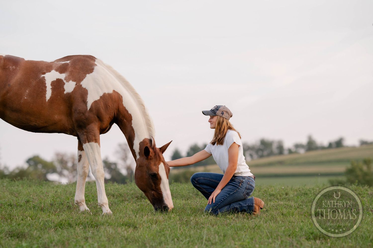 Person in baseball cap kneels beside grazing paint horse in open field.