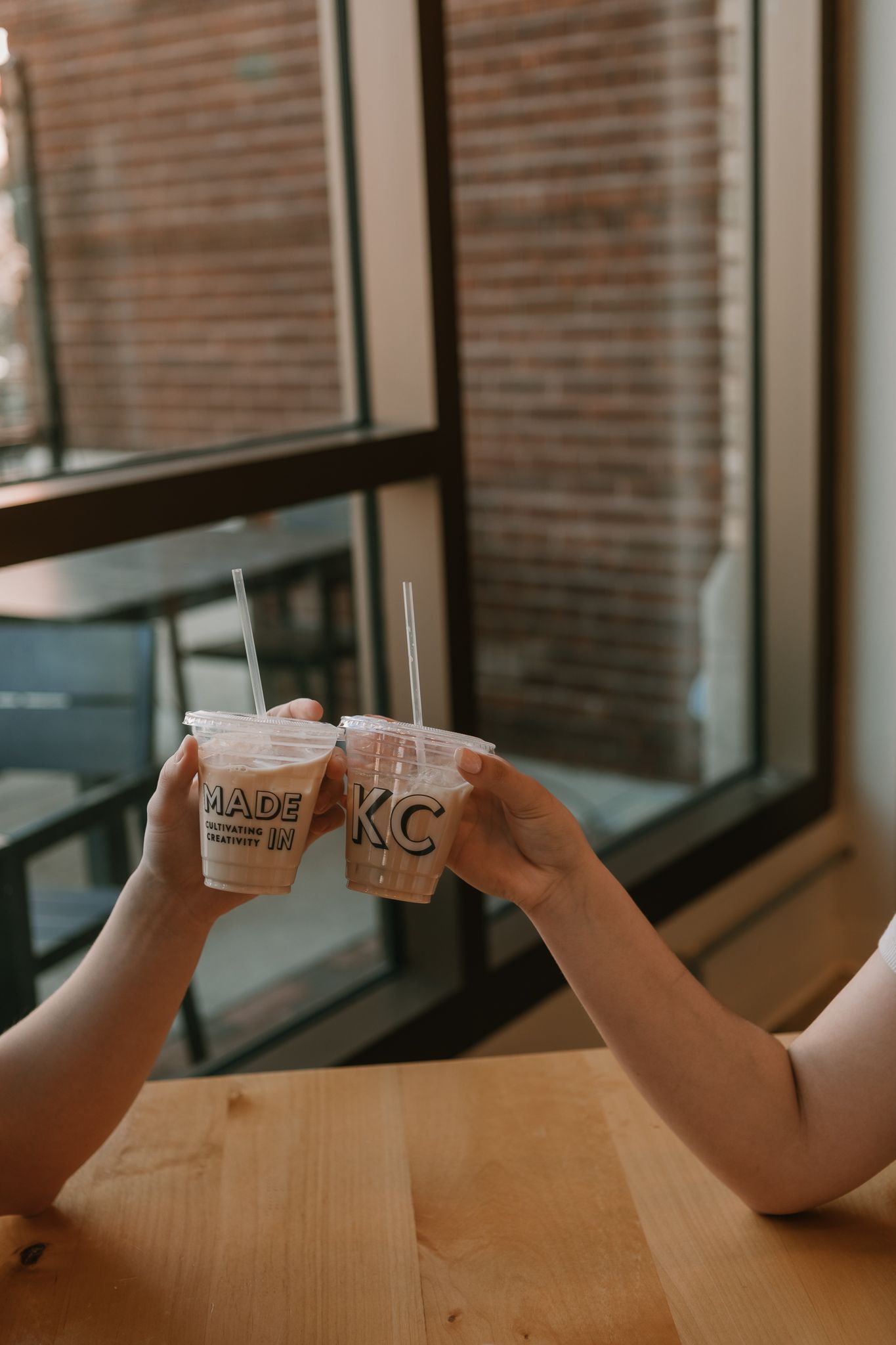 Two hands holding up plastic drinks cups for a toast at a wooden table by a window with brick wall background.