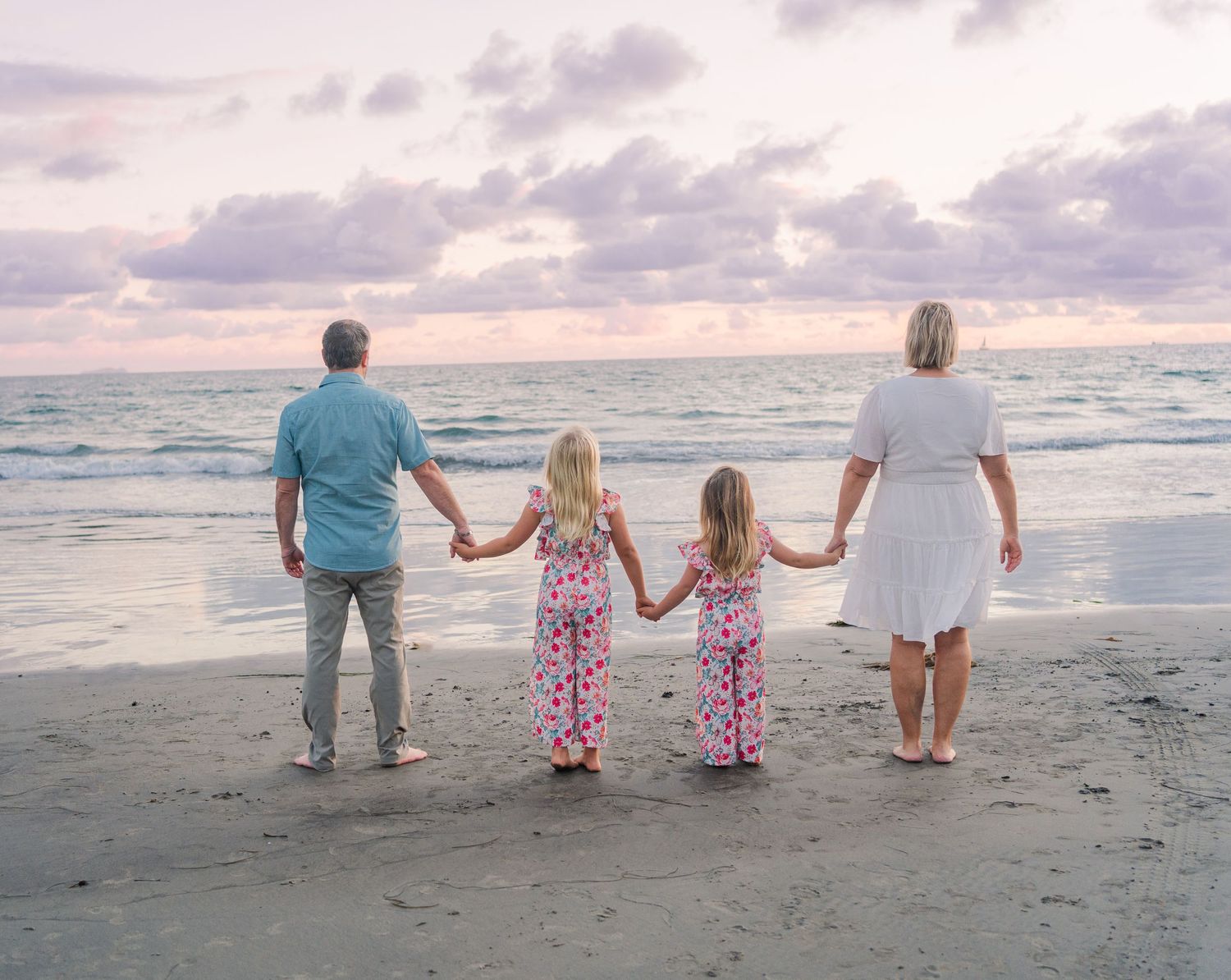 Family holding hands while facing ocean during cloudy sunset.