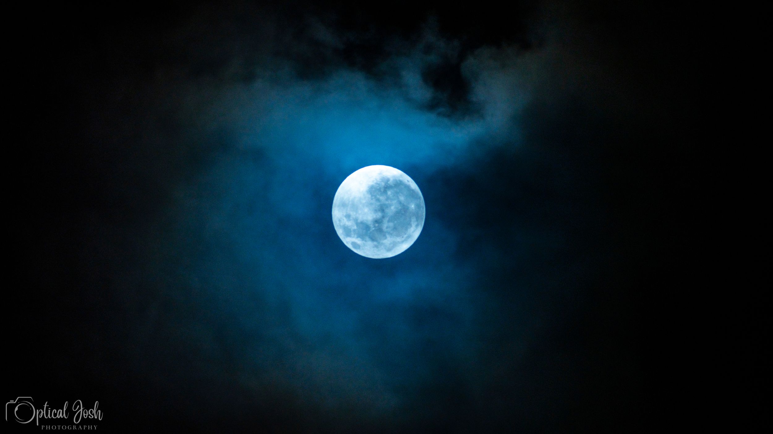 A bright full moon glowing in the dark night sky surrounded by wispy blue clouds.