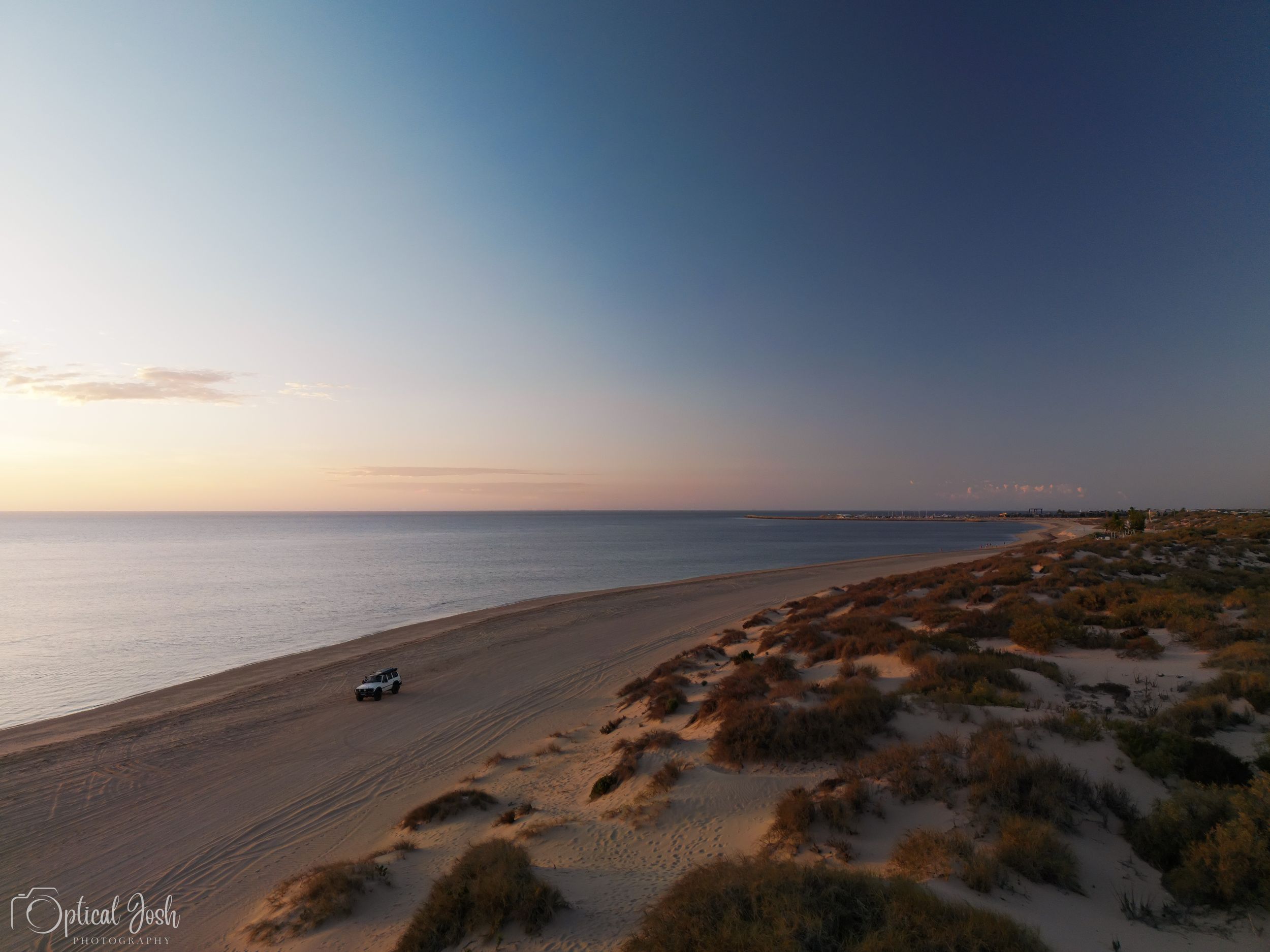 Aerial view of a serene coastline at sunset with sandy dunes and a lone vehicle parked on the empty beach.