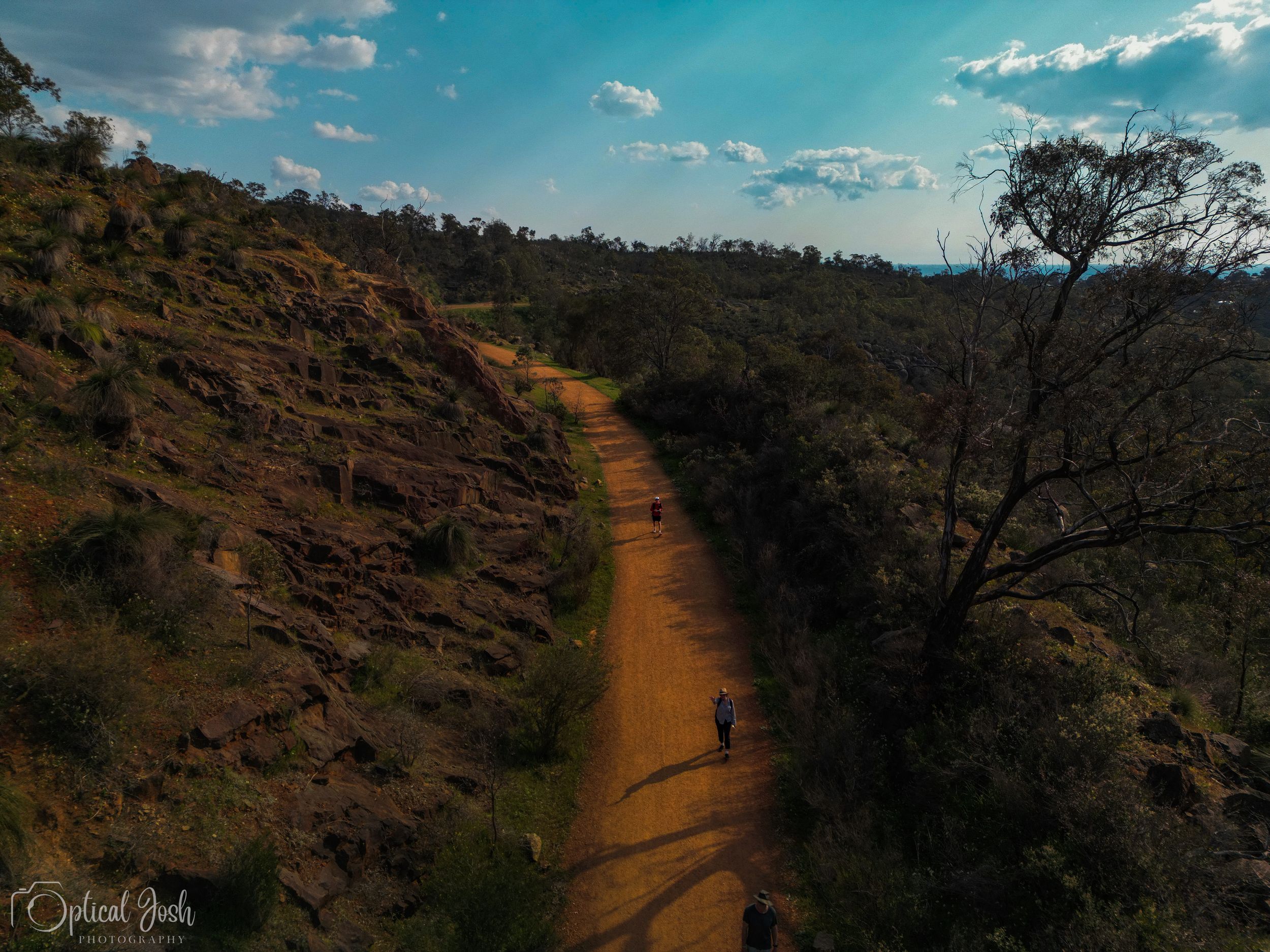 Aerial view of a winding dirt trail cutting through rugged hillside terrain under blue sky with scattered clouds.