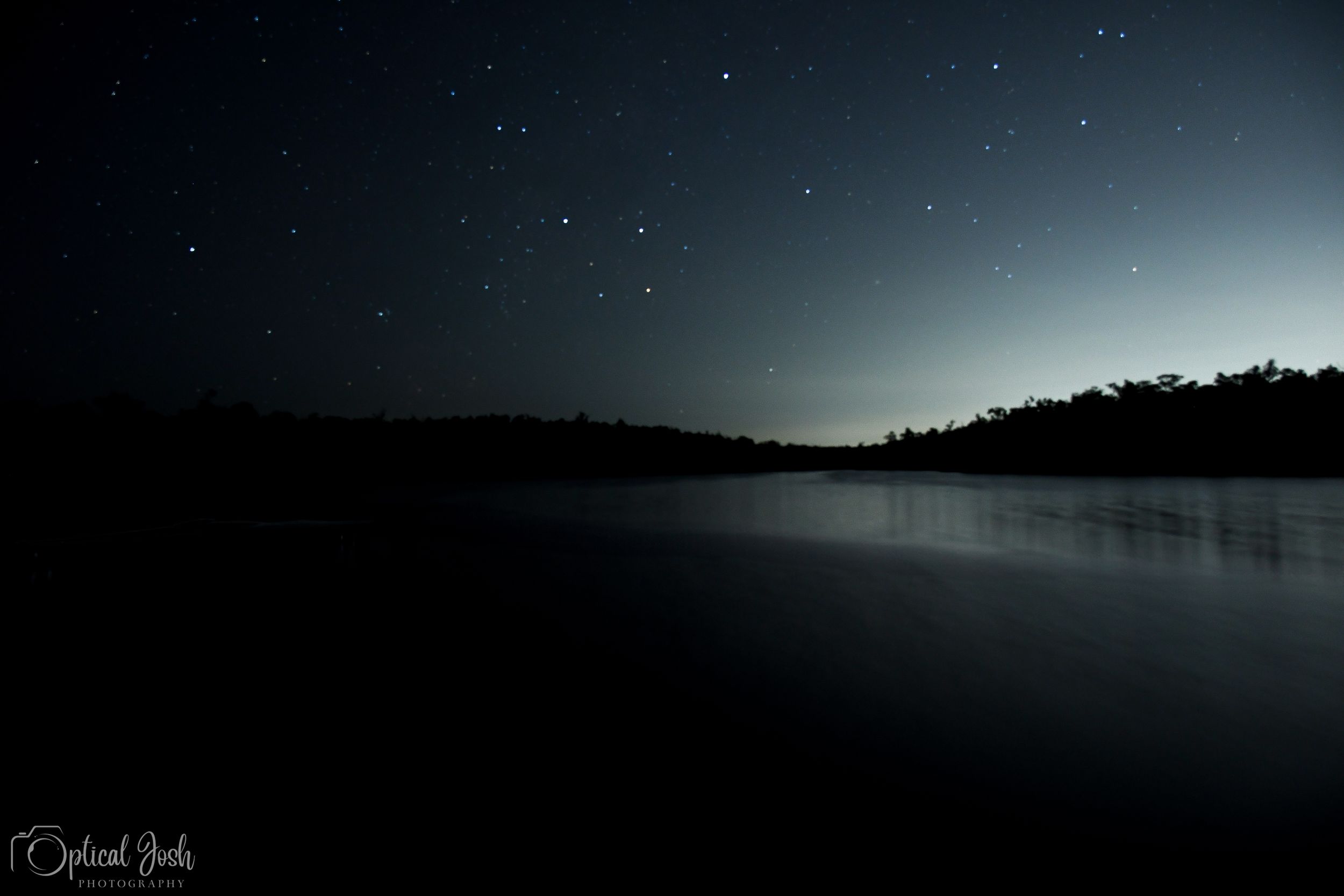 A serene nighttime lake landscape with starlit sky and silhouetted treeline reflected in calm dark waters.