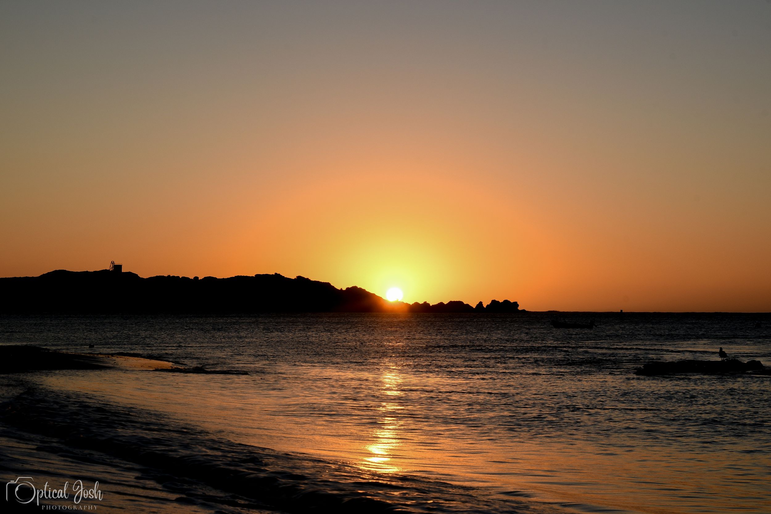 Vibrant orange sunset illuminates the ocean horizon as waves roll onto a sandy beach at dusk.