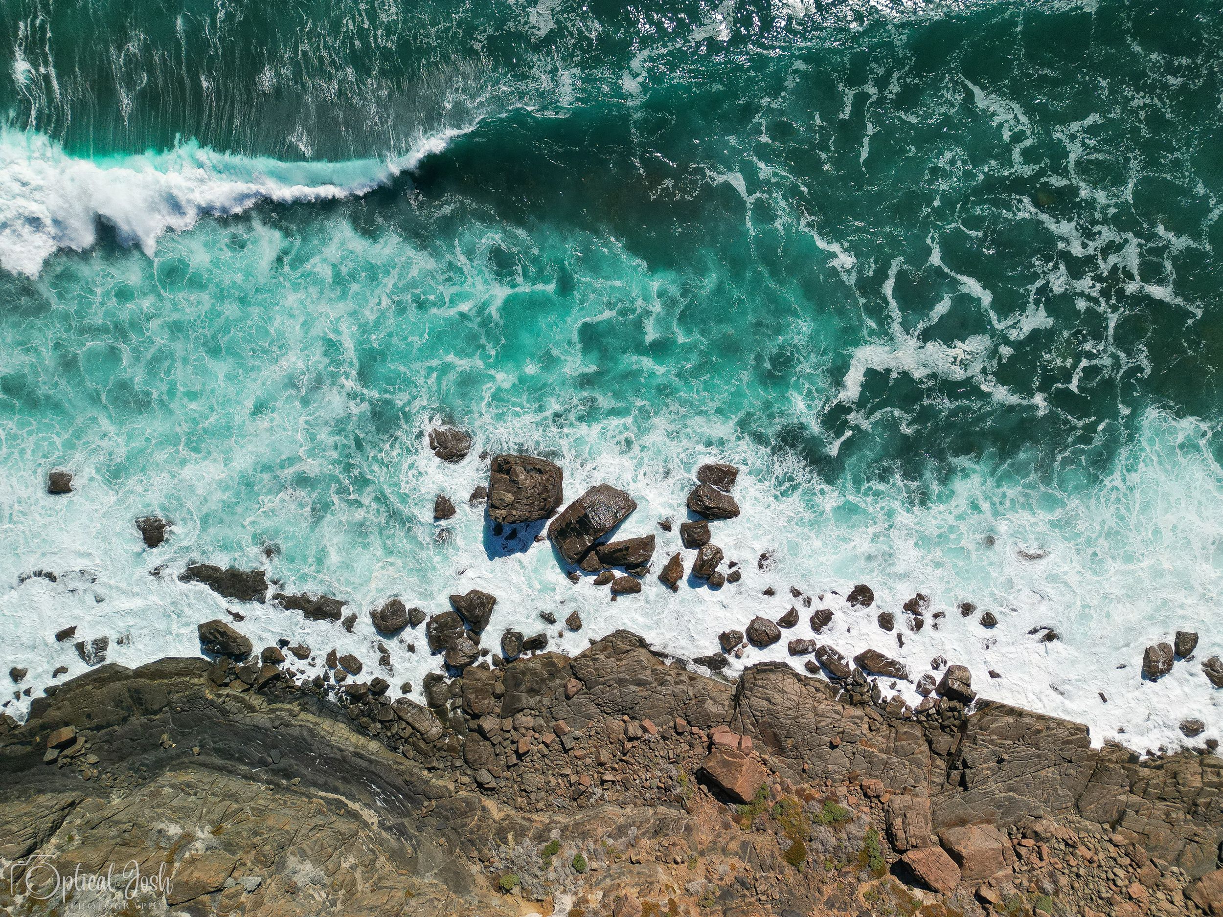 Aerial view of turquoise ocean waves crashing against dark rocky coastline from above.