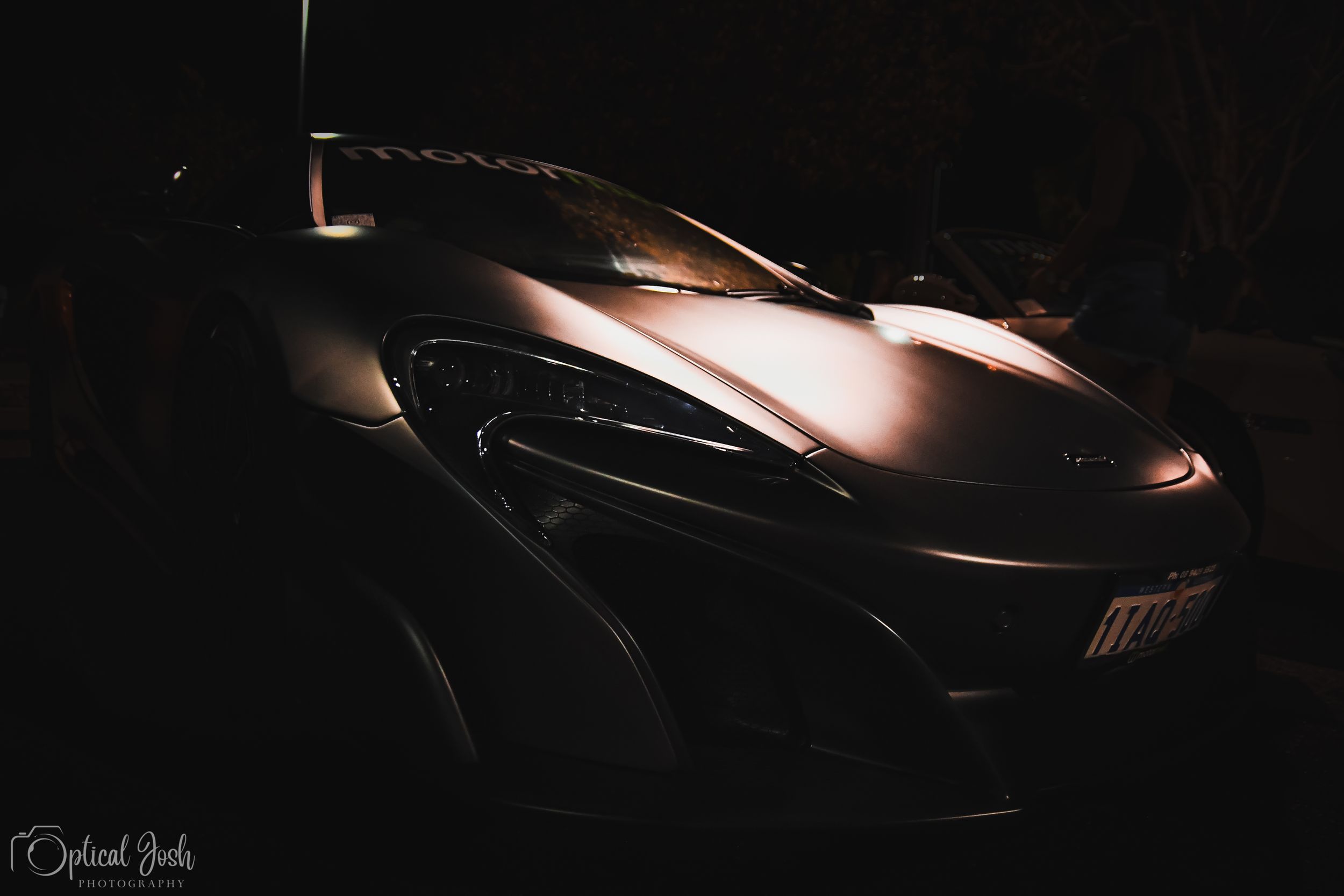 Dark artistic automotive photograph showing the front end of a luxury sports car in dramatic low-key lighting.