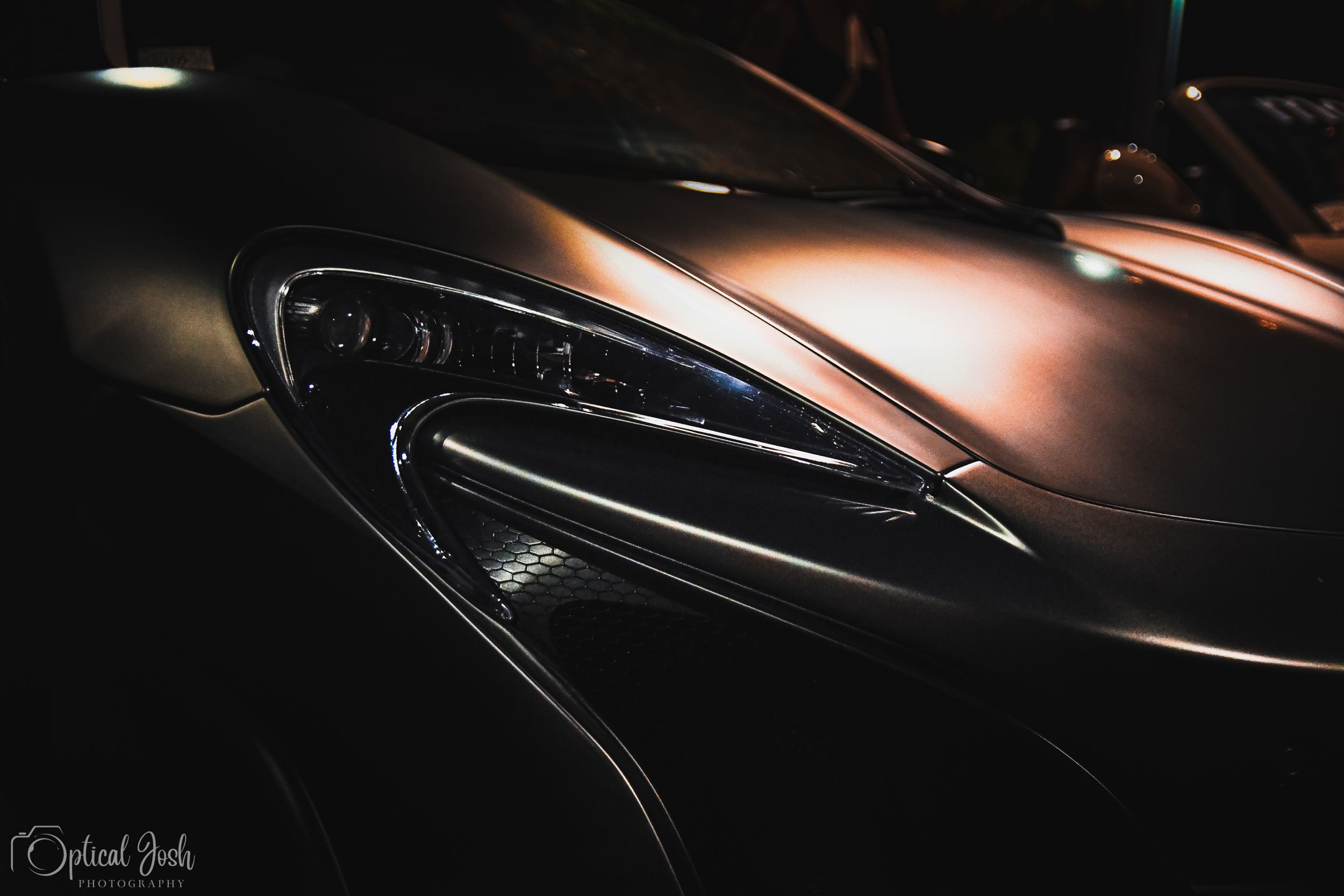 Close-up view of a sleek bronze-colored supercar headlight with curved design details in low lighting.