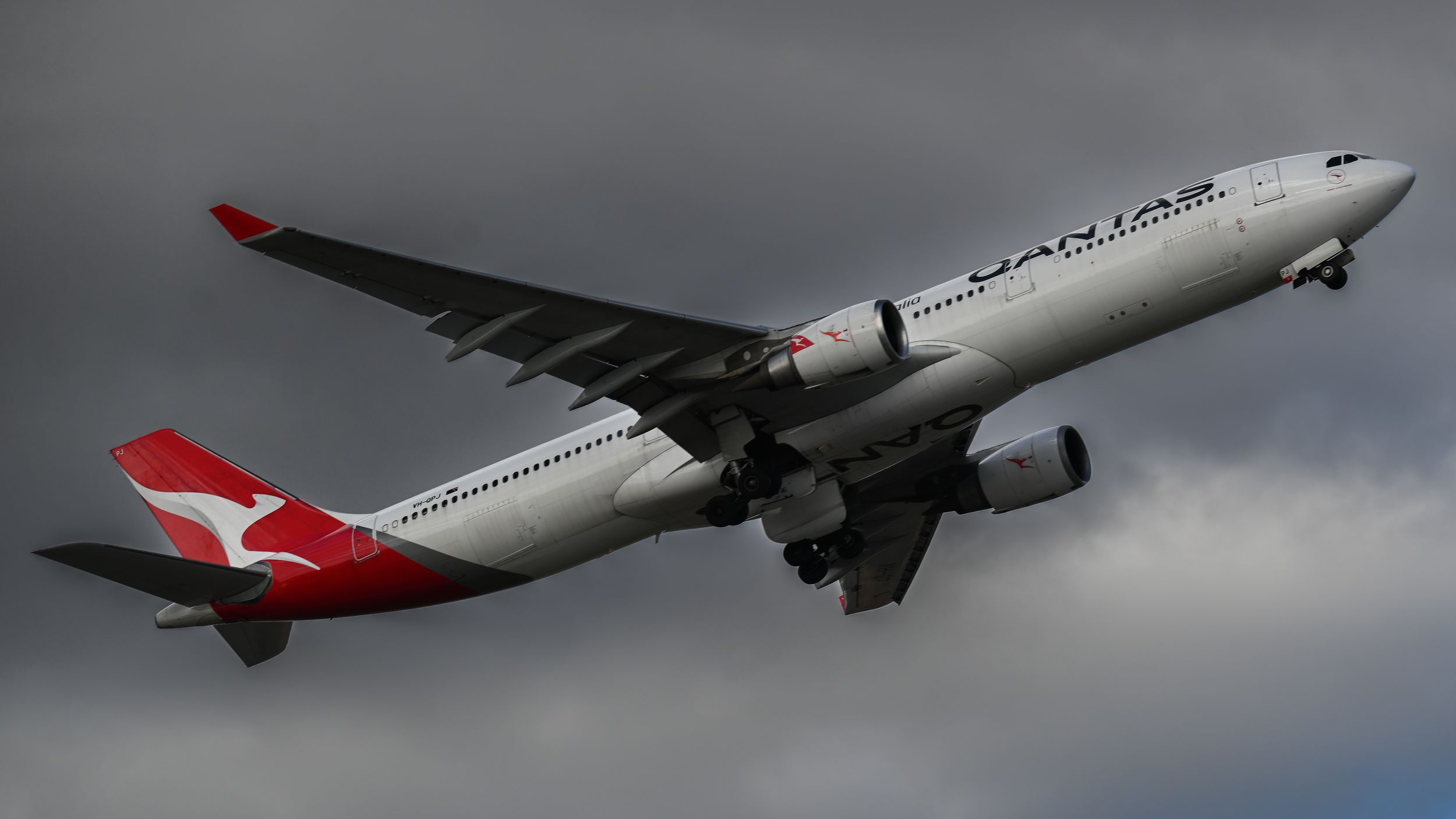 A Qantas Airbus A330 aircraft with distinctive red and white livery flying against a dark cloudy sky.