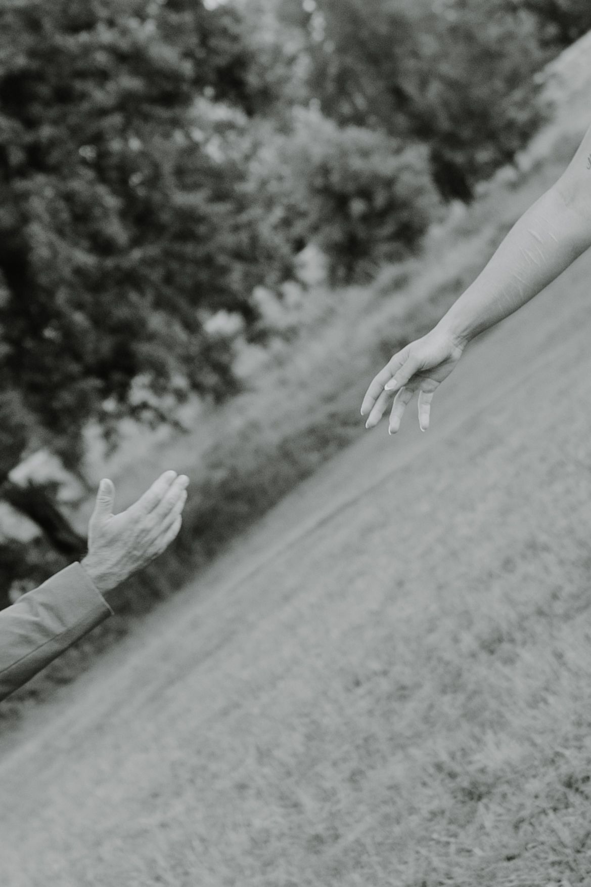 Two hands reaching towards each other against a blurred natural outdoor background in black and white photography.