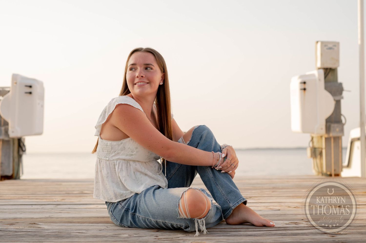 Person in white top and ripped jeans sitting on marina dock at dusk.