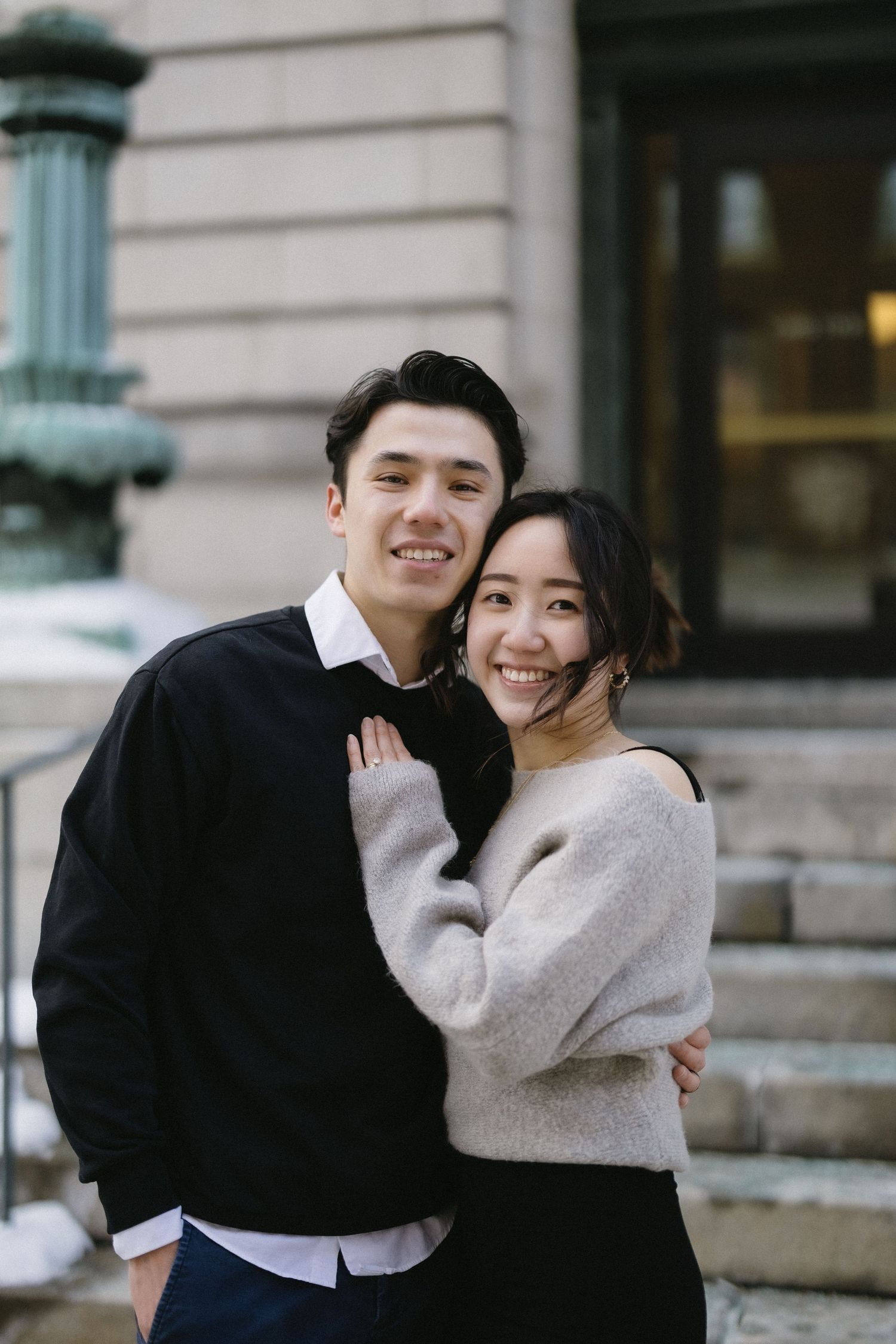 A couple embraces while smiling outdoors near stone steps wearing coordinated black and gray sweaters.