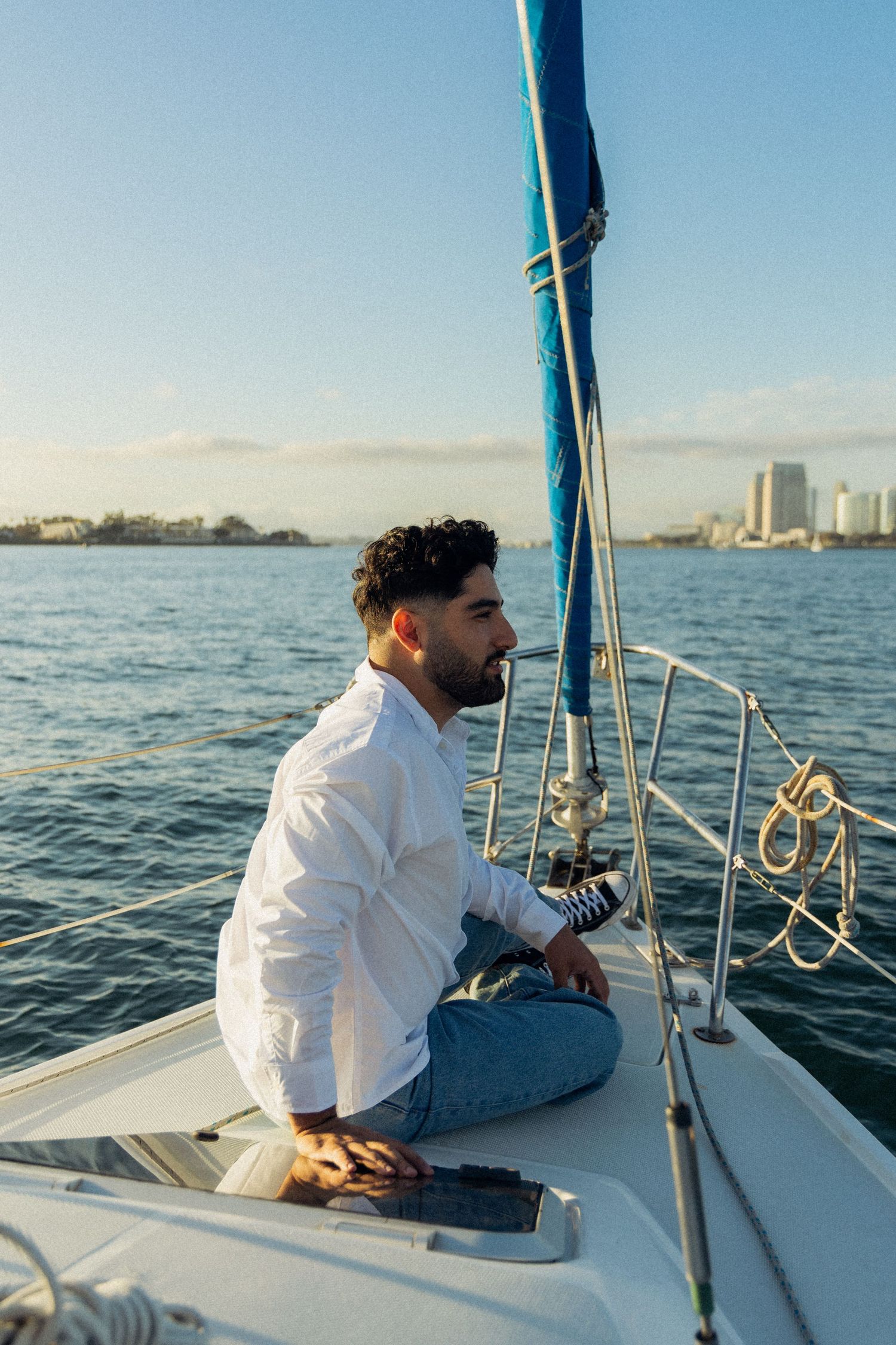 Person sailing and relaxing on a sailboat deck while cruising along a calm waterfront with a city skyline in view.