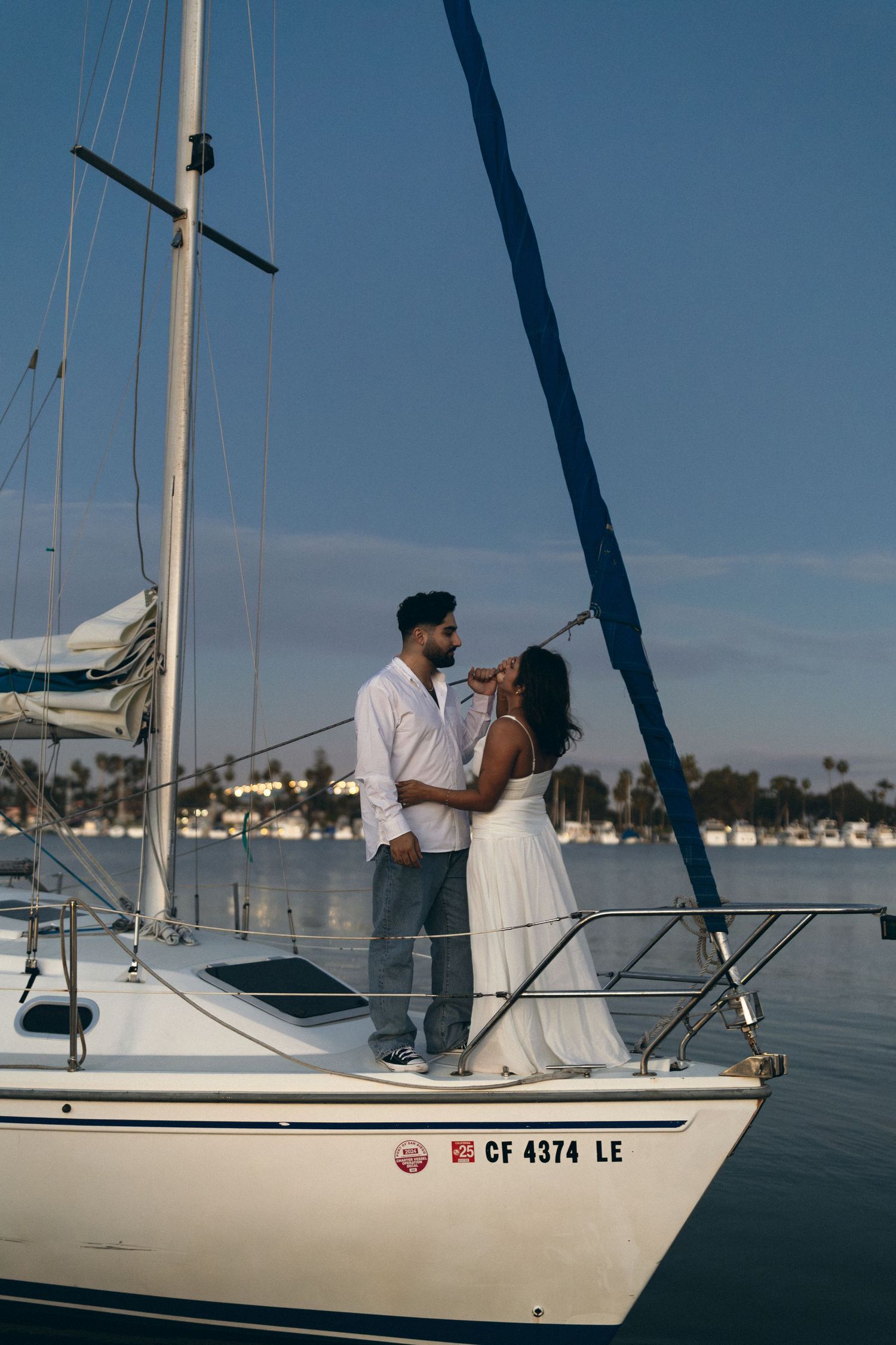 A couple shares a romantic moment aboard a sailboat docked at a marina on a clear day.