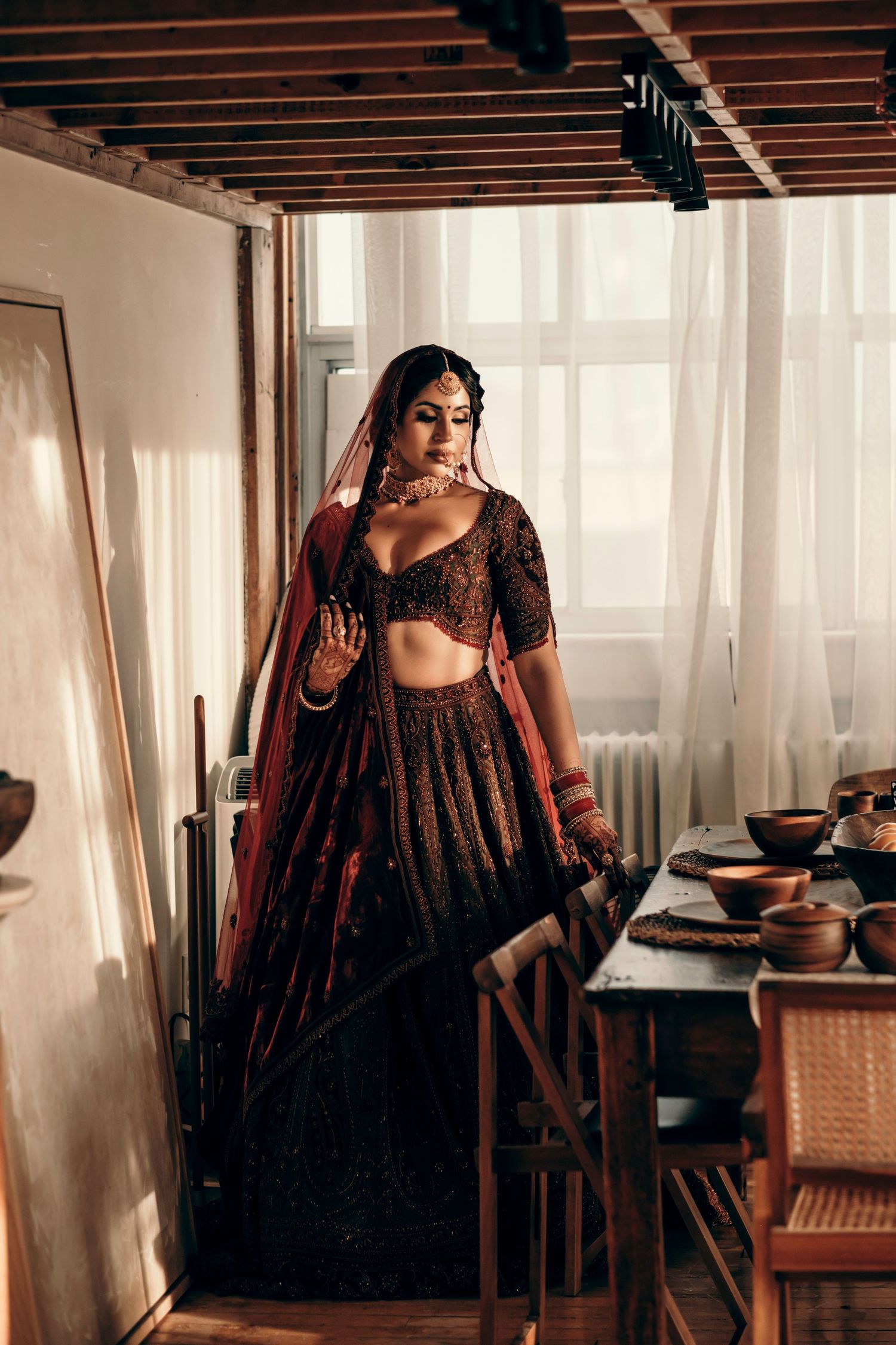 Woman wearing a traditional dark burgundy Indian lehenga with embroidered details poses elegantly in a rustic indoor setting.