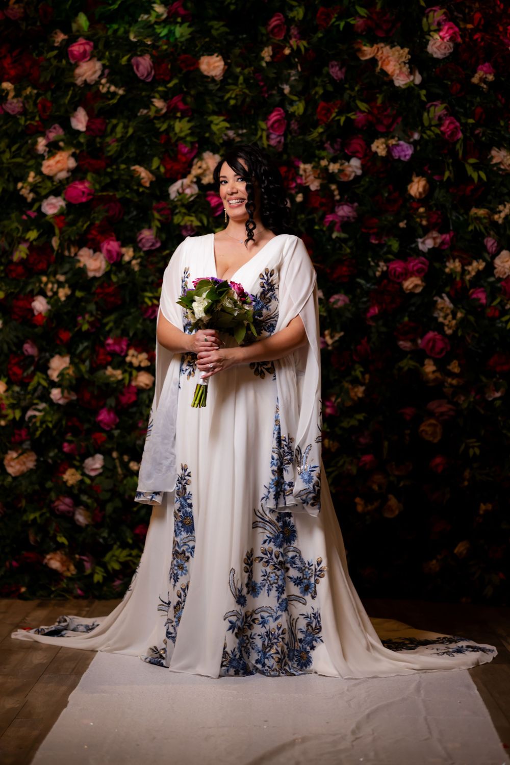 A bride in a flowing white gown with blue floral details poses against a backdrop of pink and red roses.