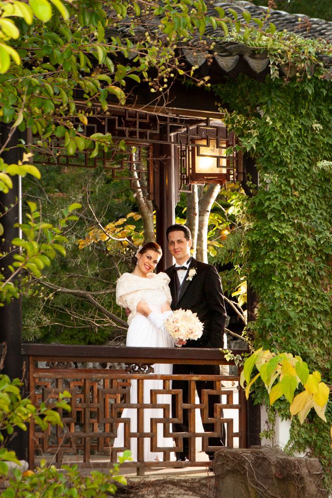 A couple poses in an ivy-covered gazebo holding a white bouquet.