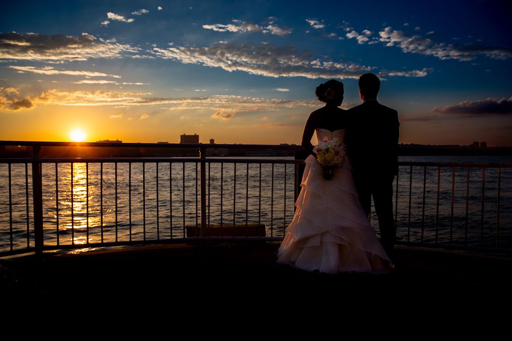 Silhouetted couple in wedding attire standing by railing at sunset over waterfront.