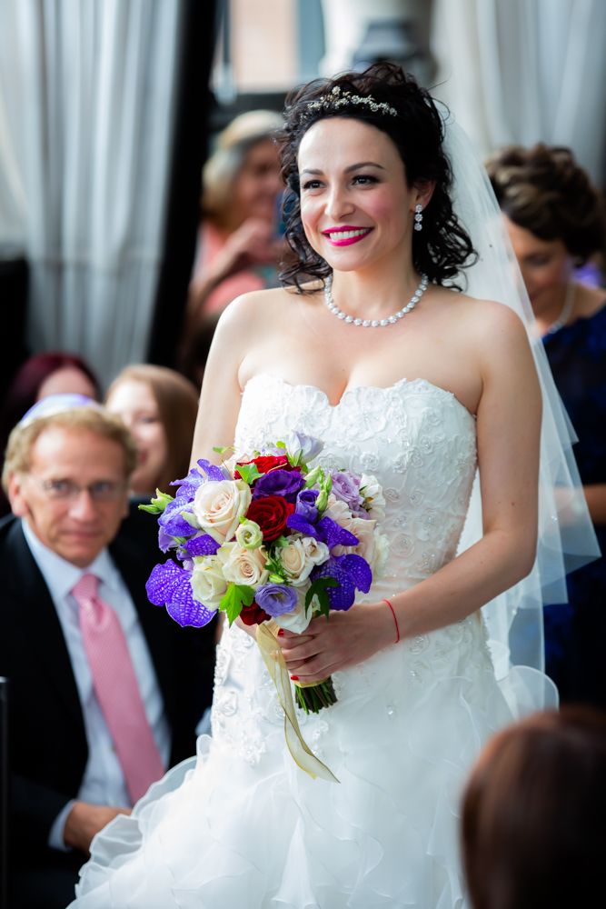 Bride holding purple and white bouquet smiles during wedding ceremony.