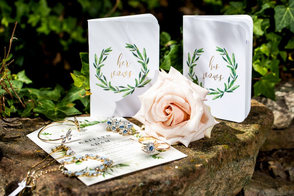 Wedding invitation cards with green leaf design alongside a pink rose and jewelry on stone surface.