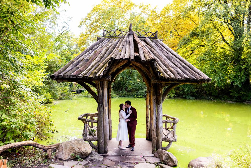 A rustic wooden gazebo with a shingled roof stands in a lush green garden setting during summer.