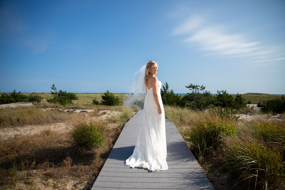 Bride in white dress walking along wooden boardwalk through coastal dunes.