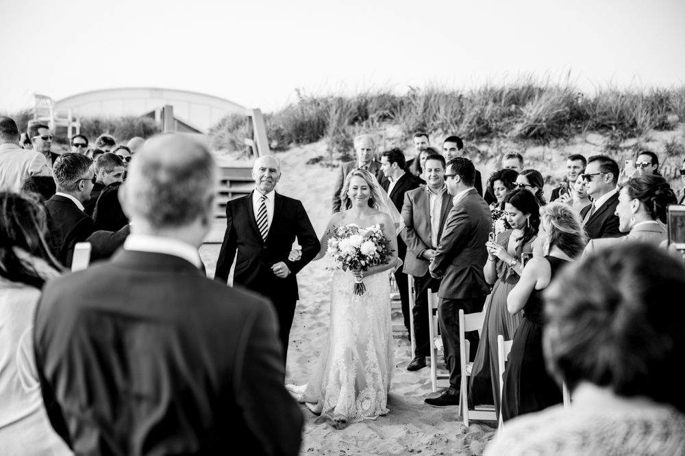 A black and white beach wedding ceremony with guests gathered on sandy dunes watching the processional.