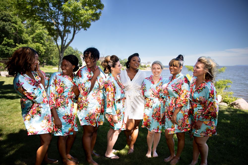 A wedding party wearing floral print robes poses together outdoors on a sunny day with water in the background.