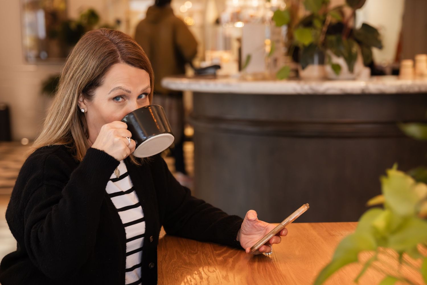 Person in a black sweater and striped shirt sits at cafe table drinking from cup while checking phone.